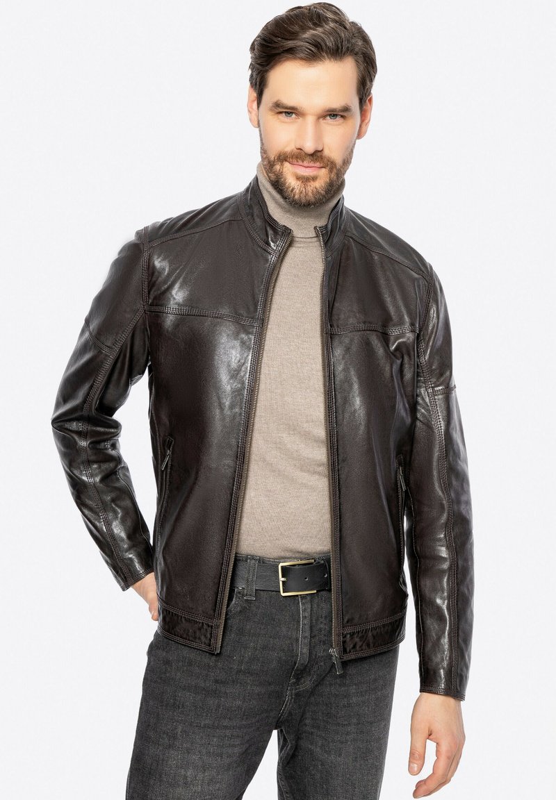Giacca di pelle marrone con zip frontale, colletto alto e texture liscia. Indossata sopra una maglia grigia chiara e abbinata a jeans scuri.