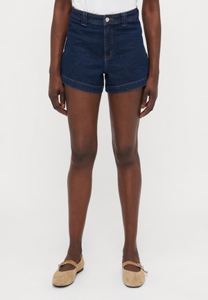 Shorts en denim bleu foncé avec une taille haute, des revers roulés et des coutures contrastantes. Associés à des ballerines beiges avec un détail de sangle.