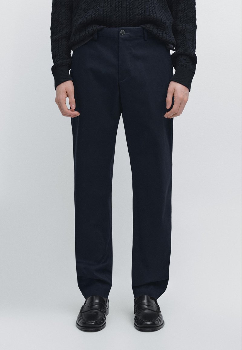 Muž nosící černé mokasíny, tmavě modré kalhoty rovného střihu a černý svetr s copánkovým vzorem stojí před jednobarevným pozadím.