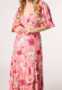 Frau in einem pinkfarbenen, blumigen Wickelkleid mit puffärmel und schillernden Rüschen, die mit gefalteten Händen vor sich steht.
