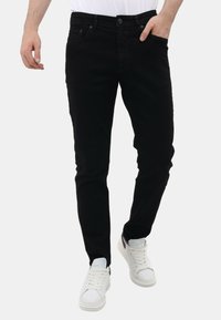 Man in slim-fit zwarte spijkerbroeken, witte sneakers en een wit shirt, met één hand in de zak en de andere hand relaxed aan zijn zijde.