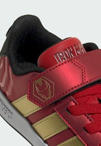 Rød og guld atletiksko med en struktureret overdel, der har en sort polstret krave, sekskantede accenter og en fremtrædende jernmaske-logo.