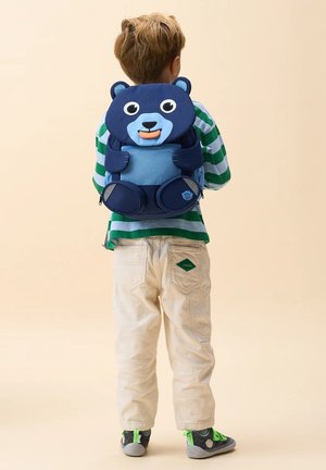Child wearing a blue bear-shaped backpack, green and white striped shirt, beige pants, and gray shoes with green accents, standing facing away.