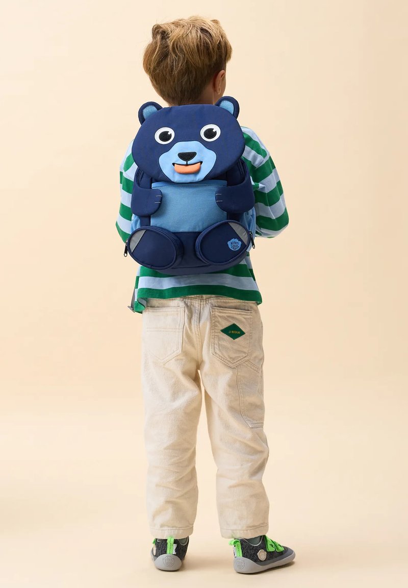 Kind mit einem blauen Rucksack in Bärenform, grün-weiß gestreiftem Hemd, beiger Hose und grauen Schuhen mit grünen Details, stehend mit dem Rücken zu.