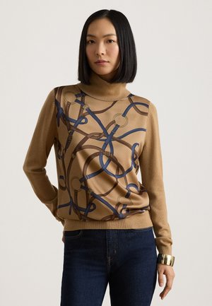 Kvinde med glat sort hår iført en brun rullekravesweater med blå og brune bæltemønstre og mørkeblå jeans, stående mod en ensfarvet baggrund.