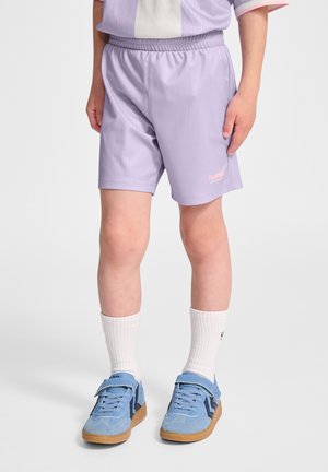 Barn iført lilla atletikshorts, hvide crew-strømper og blå sneakers, står mod en ensfarvet baggrund.