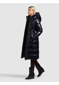 Garš tumši zils pūkains mētelis ar spīdīgu apdari, kapuci, šūtu dizainu, priekšējo rāvējslēdzēju un sānu kabatām, kopā ar melniem platformas zābakiem un melnām zeķubiksēm.