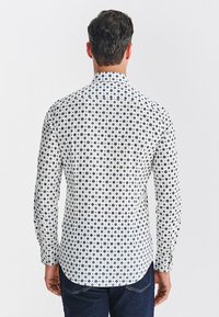 Biała koszula z długim rękawem w granatowy wzór w groszki, wykonana z miękkiej tkaniny, z zaostrzonym kołnierzykiem i zakrzywionym dołem, noszona z ciemnymi jeansami.