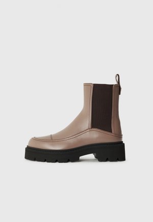 Bottines en cuir marron avec des panneaux élastiques sur les côtés, une forme arrondie à l'avant et une semelle en caoutchouc noir texturé. Dotées d'une languette à l'arrière.