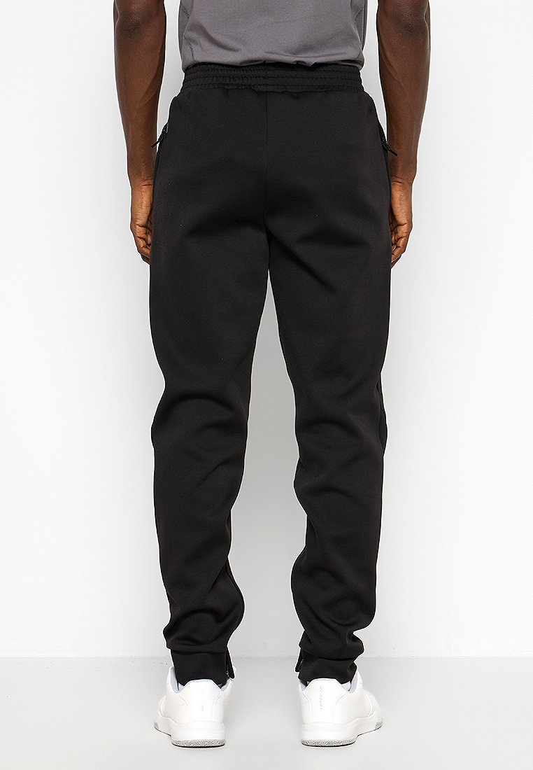 Pantalons de jogging noirs avec une ceinture élastique et des poches latérales, fabriqués en un tissu doux et texturé. Portés avec des chaussures de sport blanches.