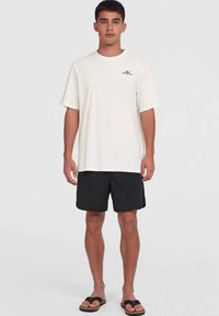 T-shirt crème clair à manches courtes avec le logo O'Neill, associé à un short noir. Le modèle porte des tongs marron sur un fond uni.