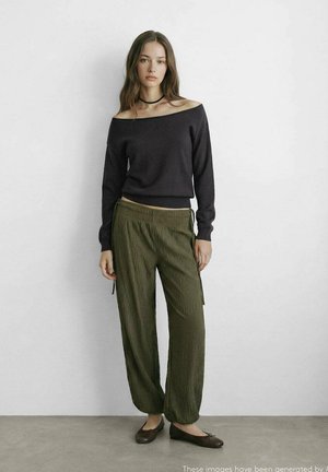 Femme debout contre un mur blanc, portant un pull noir à épaules dénudées, un pantalon texturé vert olive, des ballerines noires et un collier ras-du-cou noir.