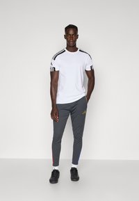 Hvid atletisk t-shirt med sorte striber på skuldrene, grå tapered bukser med røde og blå sideaccenter, sorte atletiske sko og hvide sokker.