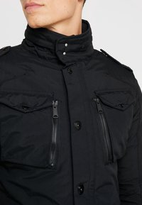 Homme portant une veste noire à col montant avec deux poches poitrine zippées et des rabats à boutons, faisant face légèrement vers la gauche.