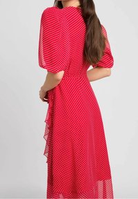 Frau mit langen braunen Haaren, die ein rotes Kleid mit weißen Punkten und Puffärmeln trägt, vor einem schlichten hellen Hintergrund stehend.