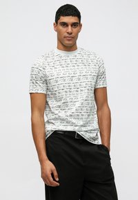 Άνδρας που φοράει λευκό κοντομάνικο T-shirt με επαναλαμβανόμενο μαύρο τύπωμα υπογραφής "KARL LAGERFELD", μαύρη ζώνη και μαύρο παντελόνι, στέκεται με το ένα χέρι στην τσέπη.