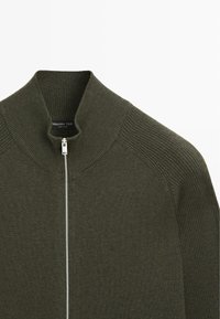 Dunkelolivgrüner Reißverschluss-Pullover mit hohem Kragen, gerippter Struktur und kontrastierendem weißen Reißverschluss. Etikett mit der Aufschrift "Massimo Dutti".
