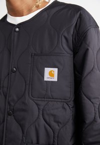 Fekete steppelt kabát hullám mintázattal, patentos záródással és mellzsebbel. Carhartt logó narancssárga színben, fehér háttér előtt.