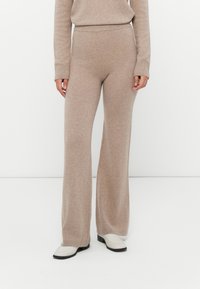 Beige gebreide wijde broek gecombineerd met een bijpassend gebreid topje en witte schoenen tegen een effen witte achtergrond.