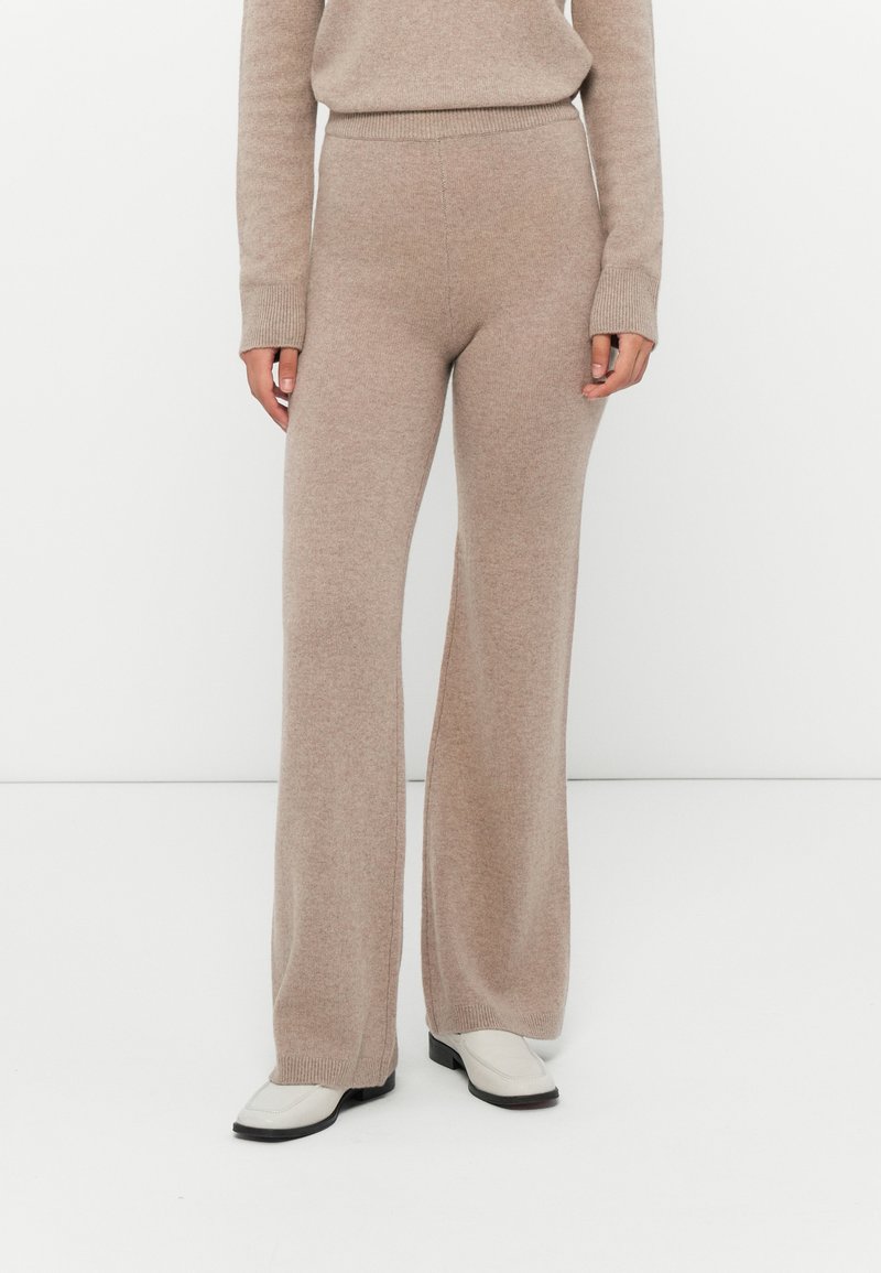 Beige gebreide wijde broek gecombineerd met een bijpassend gebreid topje en witte schoenen tegen een effen witte achtergrond.