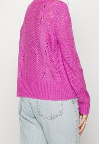 Fuchsia stickad tröja med ribbad nederkant och strukturerad mönster. Har öppna maskor för andningsförmåga, långa ärmar och en avslappnad passform.