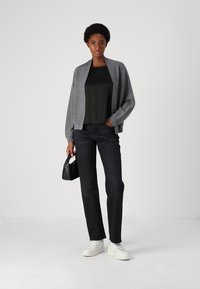 Cárdigan gris sobre una camiseta negra, combinado con jeans oscuros y zapatillas blancas. La modelo sostiene un bolso negro pequeño, de pie contra un fondo liso.