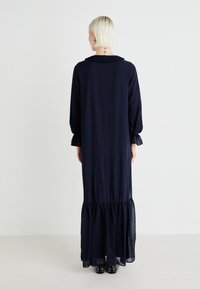 Vestido maxi azul-marinho com mangas longas, gola com volantes e detalhe translúcido texturizado na bainha, apresentando um ajuste solto e punhos elásticos.