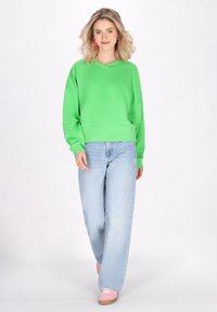 Felpa verde con scollo a V, taglio ampio e orlo a coste, abbinata a jeans a gamba larga blu chiaro e scarpe da ginnastica rosa.