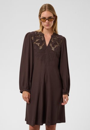 Femme portant une robe marron foncé à manches longues avec un détail en dentelle sur la poitrine et des lunettes de soleil teintées ambre, debout devant un fond uni.