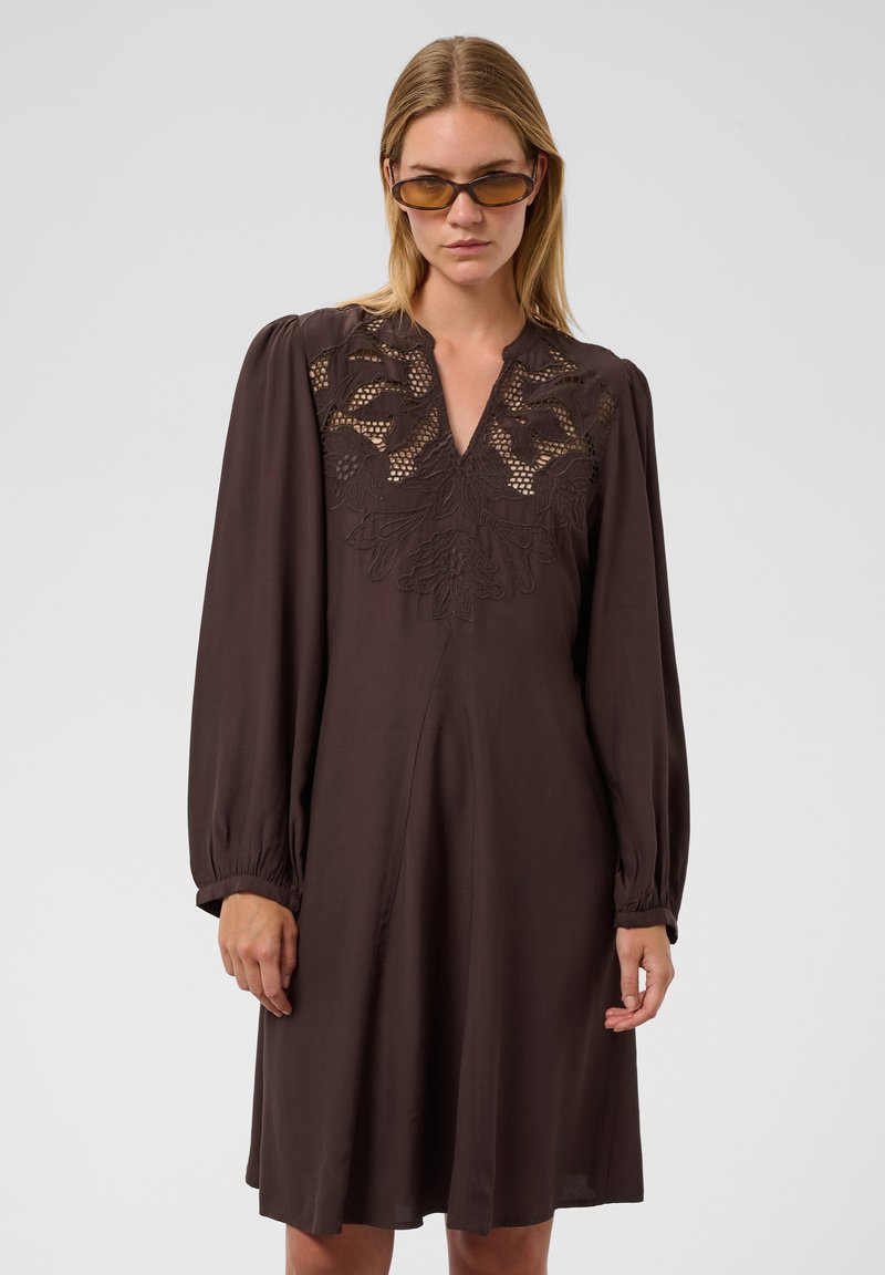 Femme portant une robe marron foncé à manches longues avec un détail en dentelle sur la poitrine et des lunettes de soleil teintées ambre, debout devant un fond uni.
