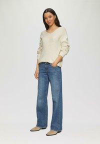 Beige, gestructureerde gebreide trui met V-hals, gecombineerd met blauwe wijde jeans. Het model draagt beige puntige schoenen. Simpele, casual outfit.
