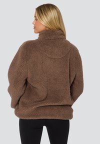Bruine fleece pullover met hoge kraag, oversized ontwerp, gestructureerd oppervlak en verlaagde schouders, gedragen over zwarte leggings.