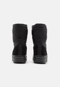 Par de botas de invierno negras con parte superior acolchada, tacón de ante y tiradores, mostradas desde atrás sobre un fondo blanco.