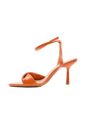 Sandalo arancione con tacco alto, punta aperta, cinturino frontale attorcigliato e sottile cinturino alla caviglia su sfondo bianco.