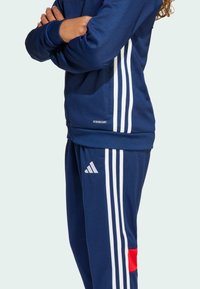 Een persoon toont een marineblauw sportoutfit met witte strepen; de top heeft een logo en de broek heeft een rode accent aan de zijkant.