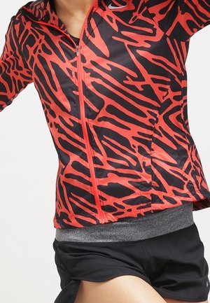 Person trägt eine rot-schwarz abstrakt gemusterte Zip-up-Jacke und schwarze Shorts, in Bewegung vor einfachem Hintergrund.