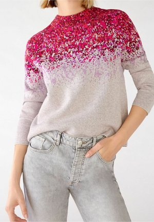 Femme portant un pull à manches longues avec un motif pixélisé dans des tons de rose et de violet, accompagné d'un jean gris clair.