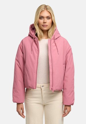 Blonde Frau, die eine rosa wattierte Kapuzenjacke über einem hellbeigen Oberteil und cremefarbenen Hosen trägt, steht vor einem einfarbigen Hintergrund.