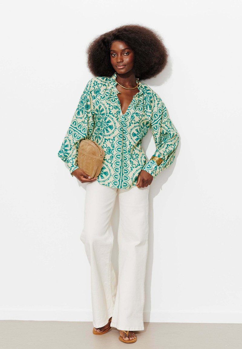 Femme avec une grande coiffure afro portant un chemisier à motifs verts, un pantalon blanc à jambes larges, tenant une pochette beige et des sandales beiges contre un mur blanc.