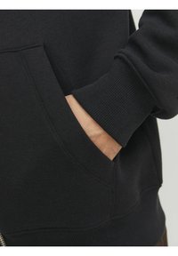 Sort sweatshirt med en sidelomme, ribbede manchetter og en blød tekstur. Nærbillede viser hånden i lommen.
