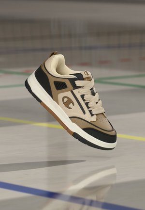 Une seule basket beige, noire et marron avec une semelle blanche épaisse, suspendue au-dessus d’un sol de gym avec des lignes de terrain colorées.
