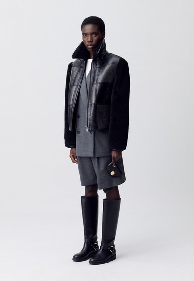 Chaqueta de borreguillo negro y cuero, pantalones cortos grises con corte a medida, y botas negras hasta la rodilla con un acento dorado. El modelo sostiene un pequeño bolso negro.