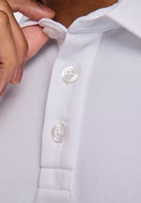 Biała koszulka polo z fakturowanej tkaniny, z trzema guzikami przy kołnierzyku oraz z górnym guzikiem z wygrawerowanym napisem "FALK". Zbliżenie na detale.