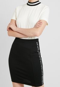 Vit kortärmad topp med svartrandig krage, tillsammans med en svart figurnära kjol med vit logotype-text på sidan. Släta texturer.