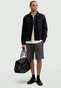 Man wearing black denim jacket, gray shorts, black shoes and sunglasses, holding large black leather bag in right hand.
