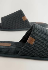 Chaussons en tissu tissé gris foncé avec un dessus texturé, un dos ouvert et une accent en cuir marron rectangulaire sur le côté. Design à semelle plate.