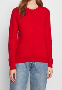 Roter Cardigan aus Strickstoff, mit langen Ärmeln, einem runden Ausschnitt und Knopfverschluss auf der Vorderseite. Getragen mit blauen Jeans.
