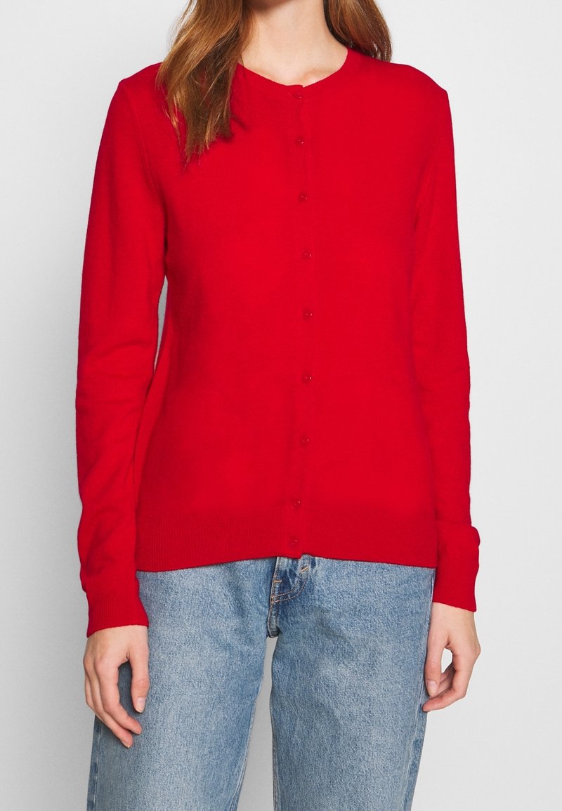 Roter Cardigan aus Strickstoff, mit langen Ärmeln, einem runden Ausschnitt und Knopfverschluss auf der Vorderseite. Getragen mit blauen Jeans.