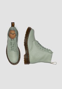 Botas de tobillo de cuero verde claro con un diseño de cordones, suelas texturizadas marrones y un acento de costura amarilla. Incluyen una pestaña para tirar.