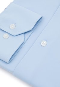 Jasnoniebieska koszula z długim rękawem z zapięciem na guziki, o gładkiej fakturze materiału i białych plastikowych guzikach. Starannie złożona, ukazująca prostotę w designie.